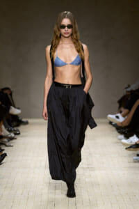 Model on a runway wearing a blue glittery triangle bikini top, black suspender straps, and loose black trousers with a belt, plus sunglasses.