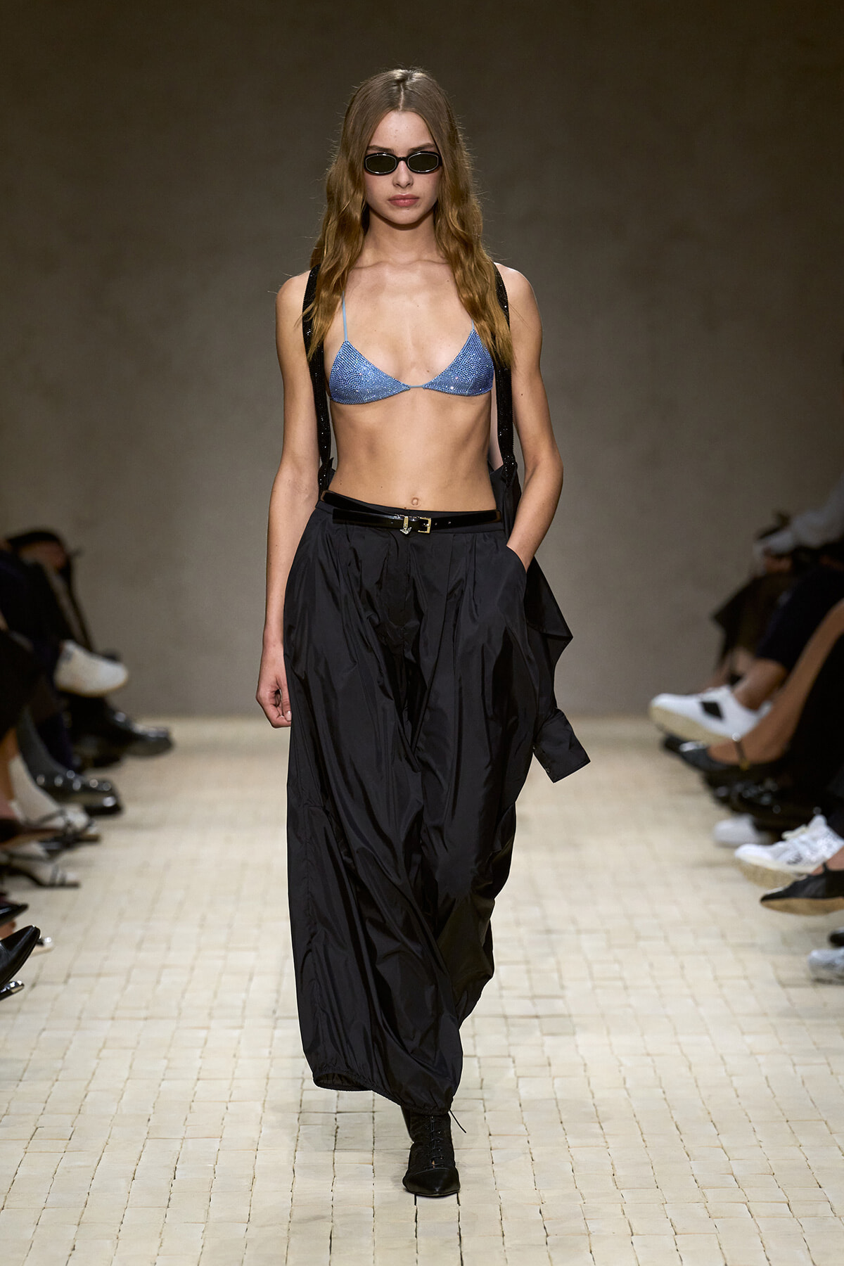 Model on a runway wearing a blue glittery triangle bikini top, black suspender straps, and loose black trousers with a belt, plus sunglasses.