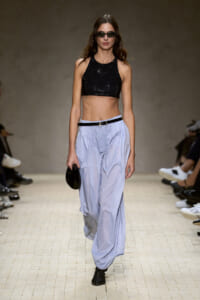 Model on a runway wearing a black sequined crop top, light blue baggy pants, black belt, sunglasses, and a small black clutch.