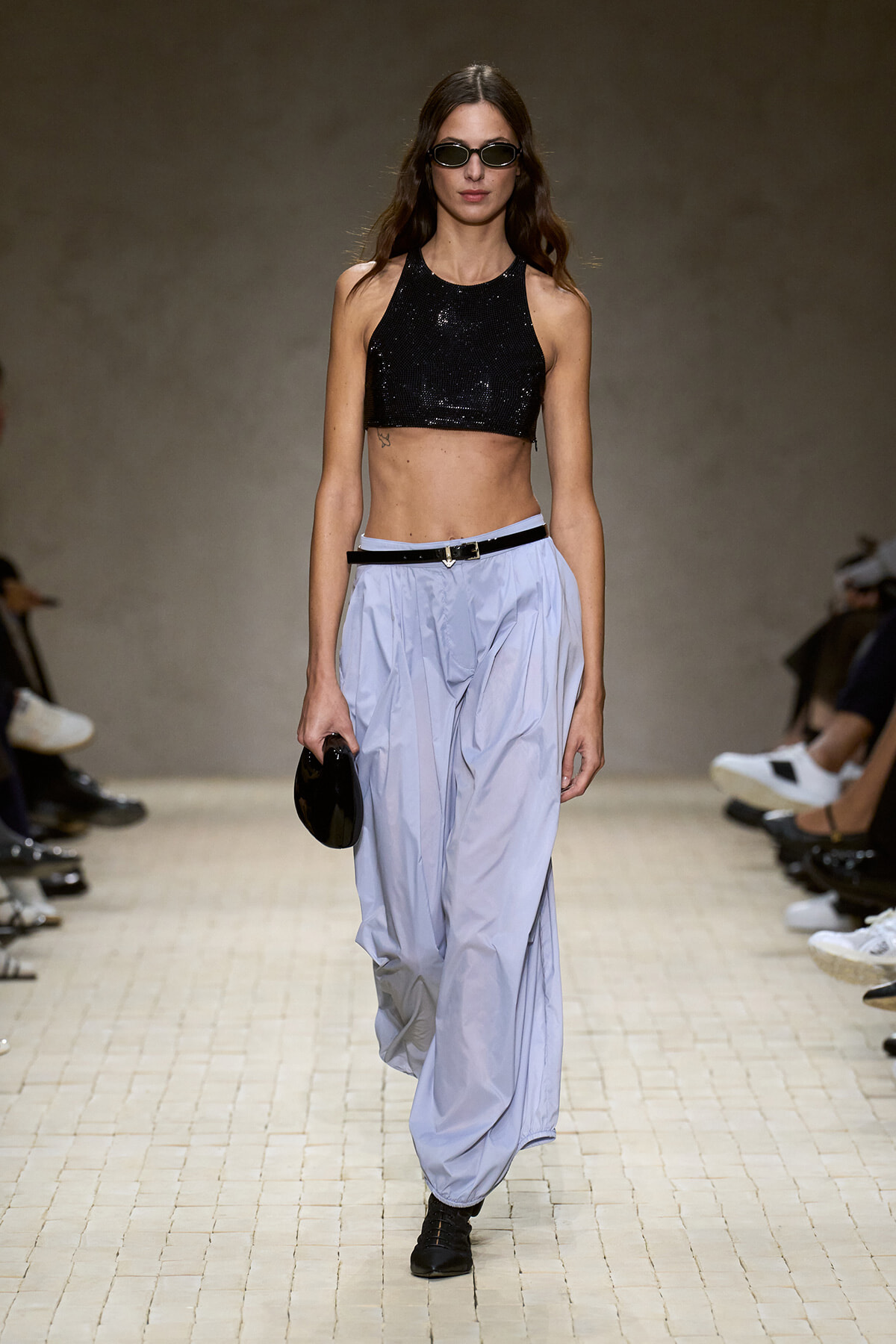 Model on a runway wearing a black sequined crop top, light blue baggy pants, black belt, sunglasses, and a small black clutch.