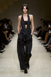 Model walks the runway in a black halter jumpsuit with a draped front and wide, voluminous pants; hands in pockets during a fashion show