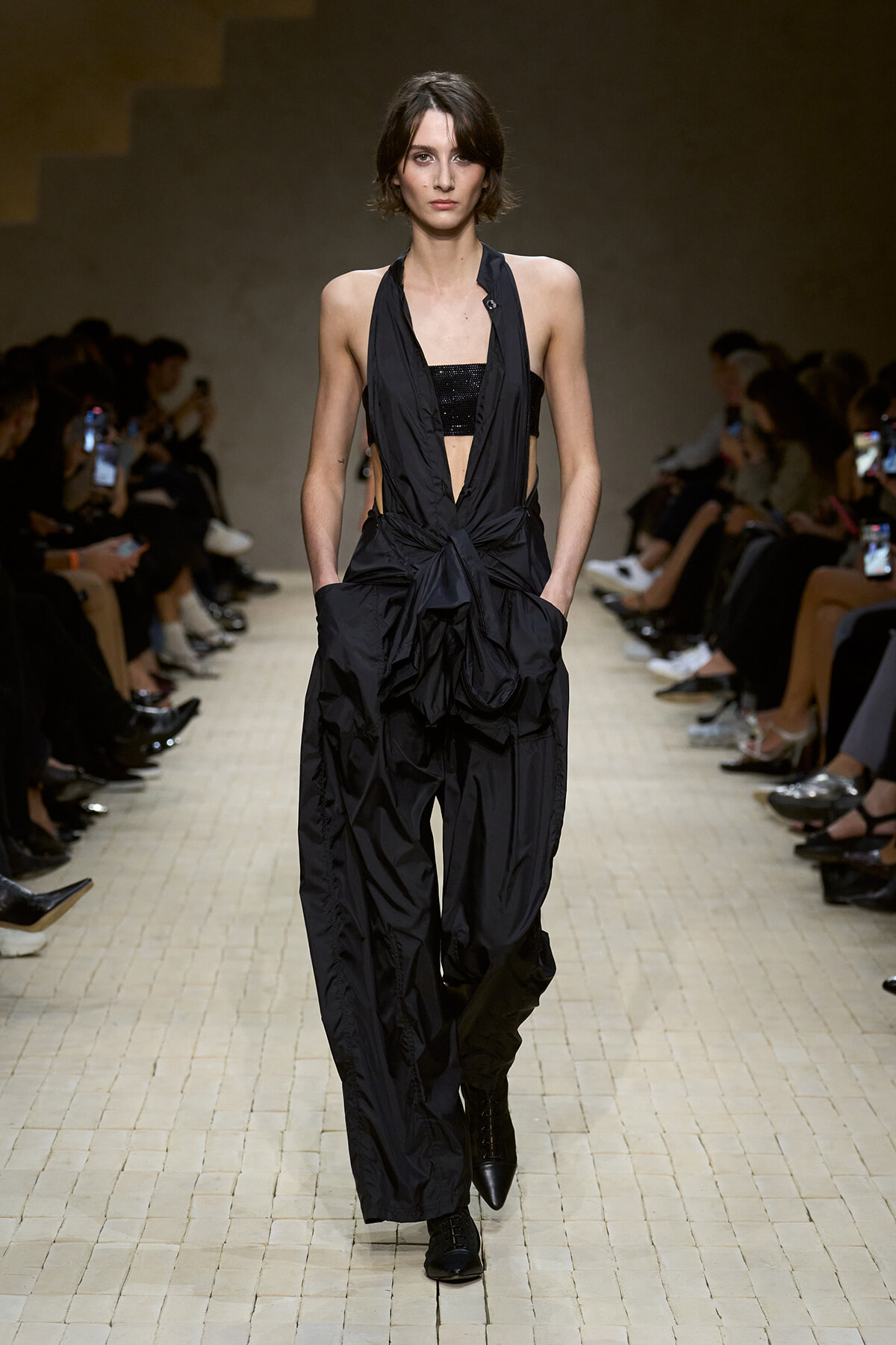 Model walks the runway in a black halter jumpsuit with a draped front and wide, voluminous pants; hands in pockets during a fashion show