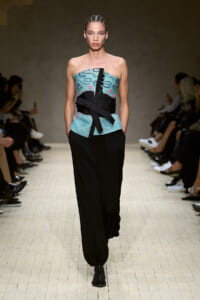 Model walks the runway in a strapless teal top with a bold black obi-style belt and loose black pants; braided hair.