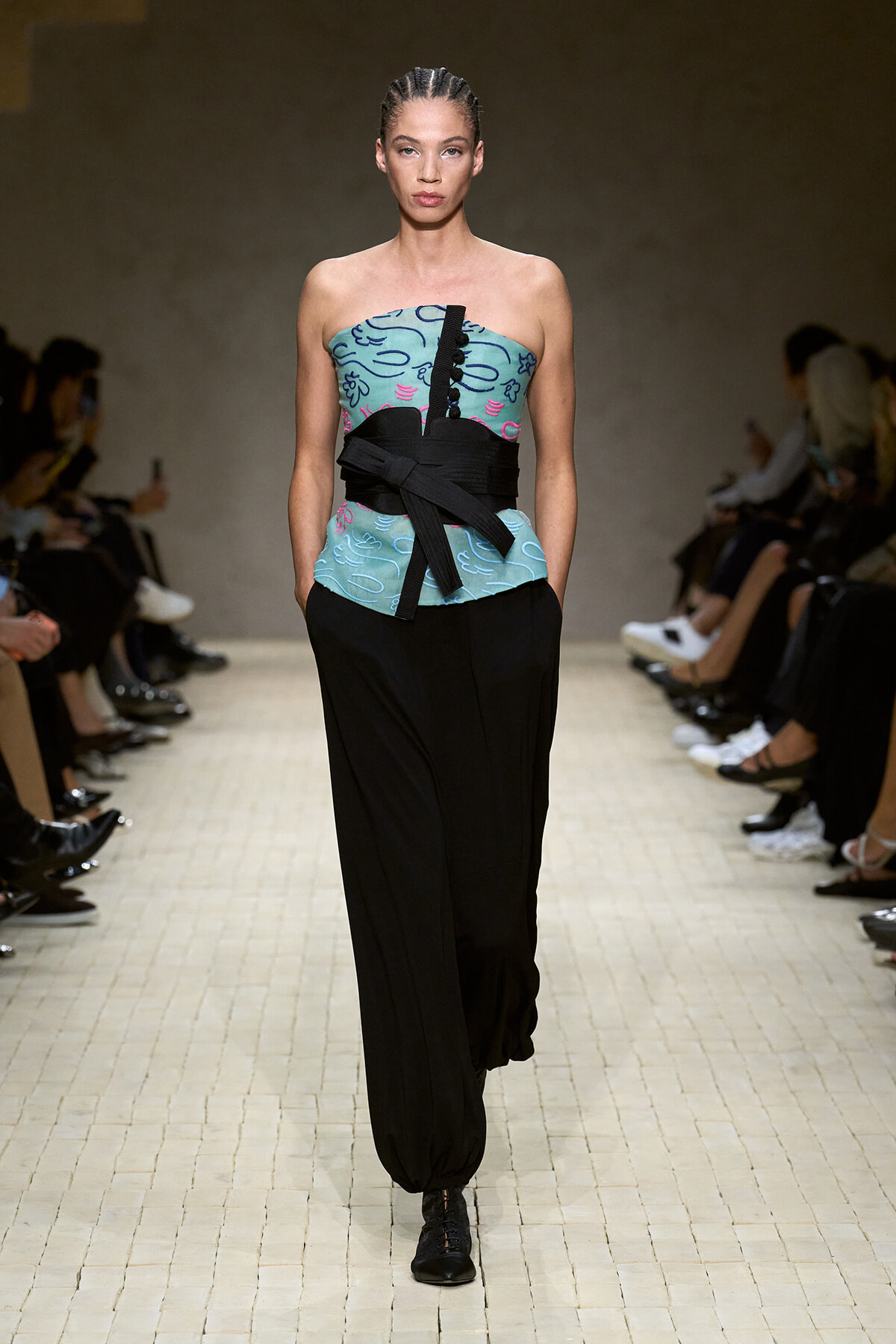 Model walks the runway in a strapless teal top with a bold black obi-style belt and loose black pants; braided hair.