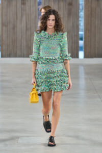Model walking in a green multicolor ruffled dress, holding a yellow handbag outdoors.