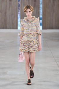 Model walking runway in a pastel tweed ruffled dress with pink handbag and brown sandals, showcasing a high-fashion look.