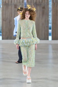 Model walks down runway in pastel green knit set with matching cropped cardigan and midi skirt, and a feathered fringe around the hips; yellow leaf headpiece adds whimsy.