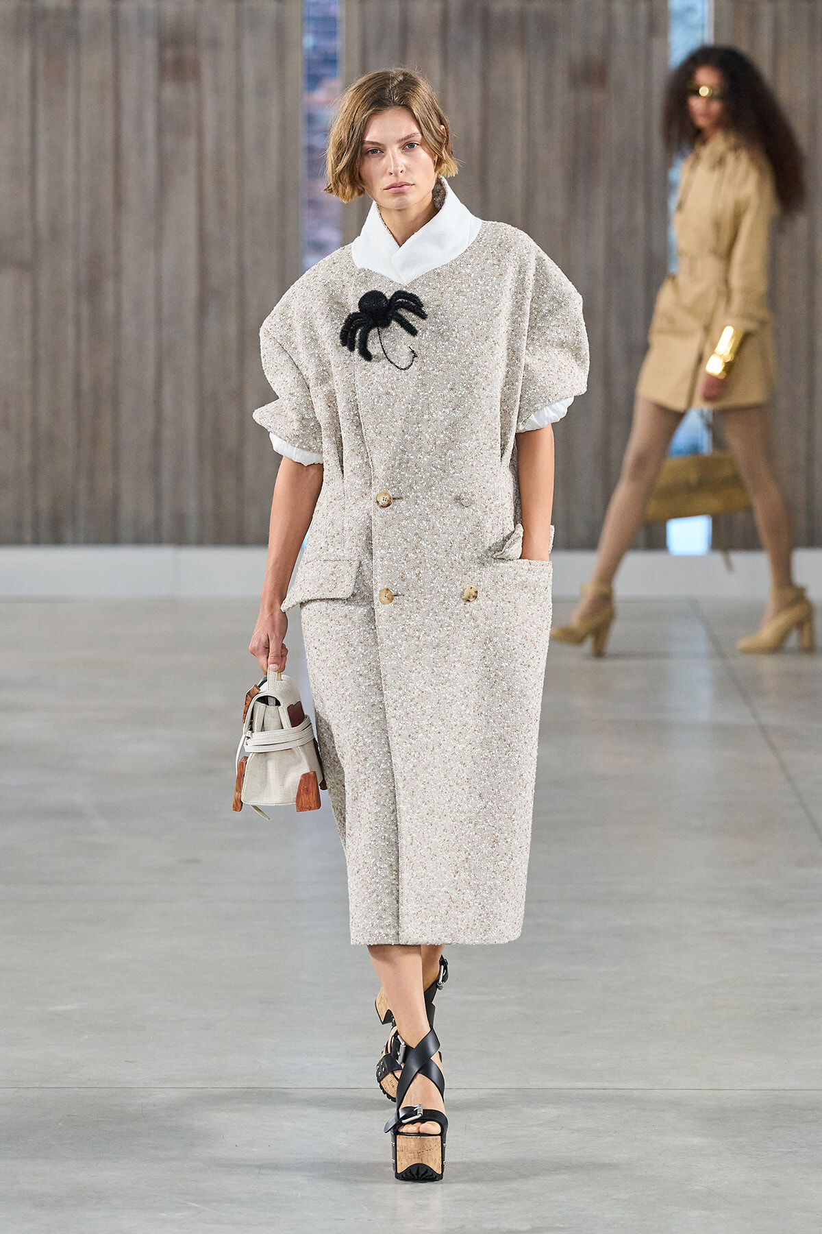 Model walks the runway in an oversized beige textured suit with a white collar, black spider brooch, and matching skirt; she carries a small handbag.