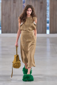 Model walking a runway in a tan jumpsuit with ruffle sleeves, carrying a mustard bag, and wearing bright green fluffy platform shoes.