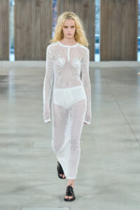 Female model walks a runway in a white mesh long-sleeve jumpsuit, revealing white undergarments, wearing black open-toed shoes in a minimalist setting.