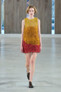 Model walking a runway in a sleeveless, gradient knit dress (yellow to orange to red) with dark brown oxford shoes in a minimalist venue.
