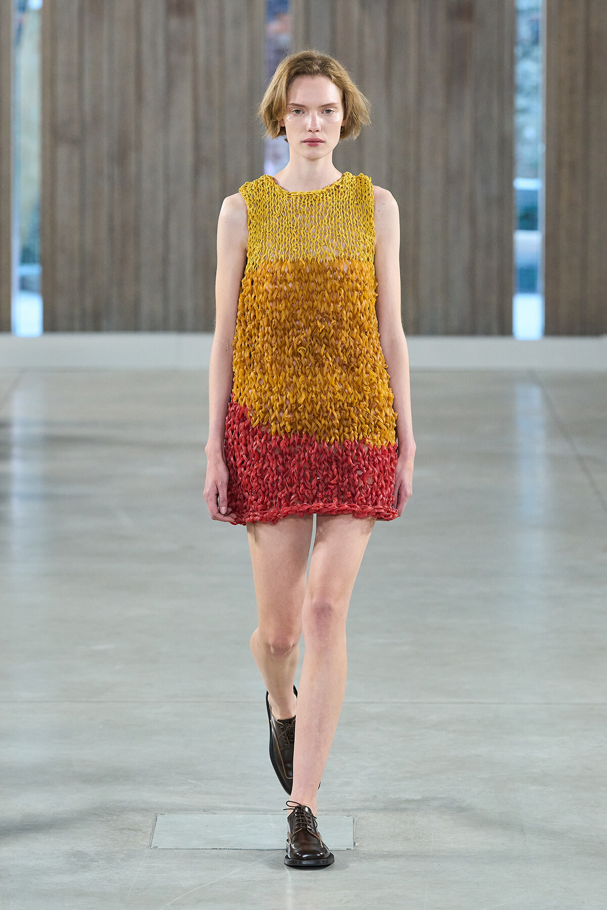Model walking a runway in a sleeveless, gradient knit dress (yellow to orange to red) with dark brown oxford shoes in a minimalist venue.