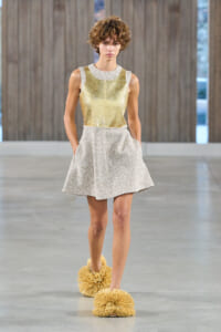 Model walking a runway in a metallic gold sleeveless top and shimmering short silver skirt, paired with oversized fluffy yellow footwear.