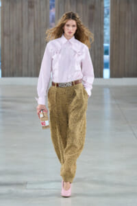 Female model walking a runway in a pale pink button-down shirt with bow details, textured tan pants, pink heels, and a Moschino belt, holding a carton prop.