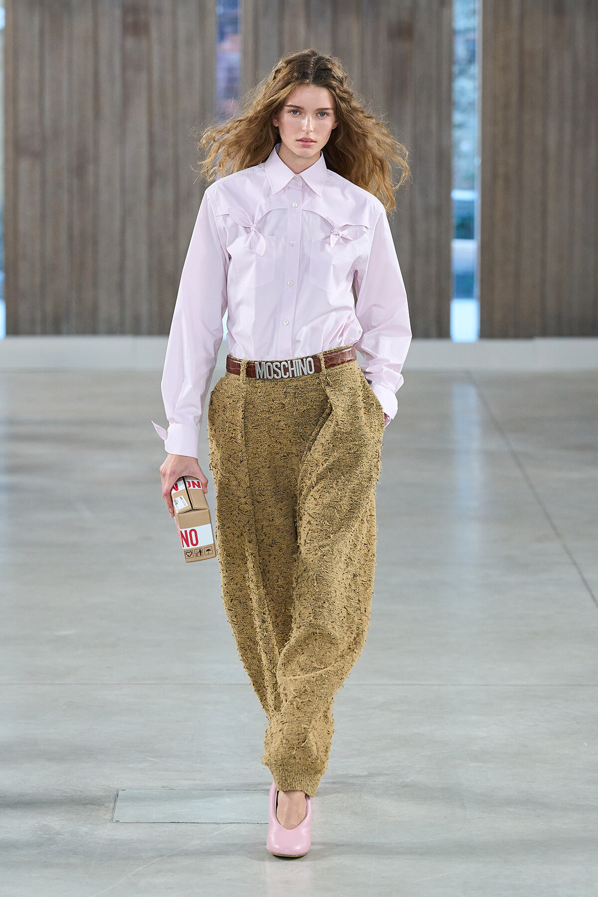 Female model walking a runway in a pale pink button-down shirt with bow details, textured tan pants, pink heels, and a Moschino belt, holding a carton prop.