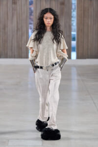 Model walks the runway in a beige ensemble with cape-style ruffled sleeves, light trousers, metallic arm cuffs, and oversized black fluffy shoes.