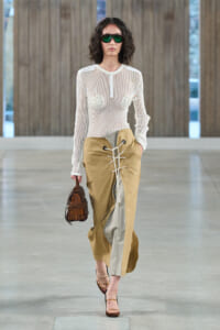 Model walking a runway in a sheer white mesh top, tan lace-up midi skirt with a gray panel, and a brown woven handbag with green sunglasses.