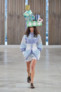 Model on a runway balancing a tall stack of colorful gift boxes on her head, wearing a blue striped shirt-dress and brown sandals for a fashion show.