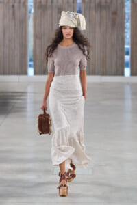 Model walks a neutral-toned runway in a beige knit top, long gray skirt, and tan platform sandals, with a cream headwrap and brown handbag.