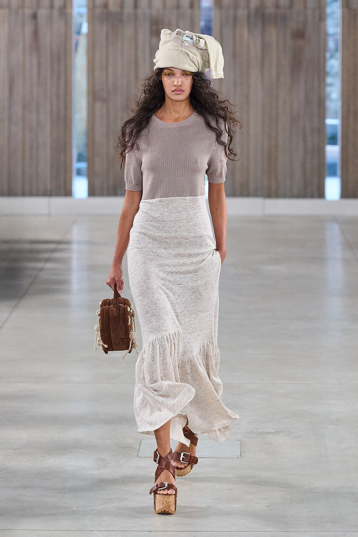 Model walks a neutral-toned runway in a beige knit top, long gray skirt, and tan platform sandals, with a cream headwrap and brown handbag.