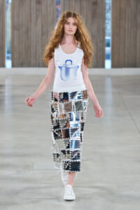 Model walking runway wearing a white tank top with a blue pot graphic and pants made of mirrored panels connected by white lattice stitching.