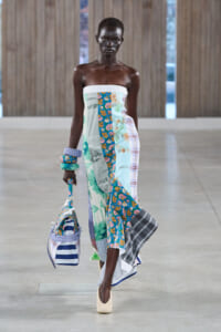 Model walking in a strapless patchwork dress with floral and plaid panels, carrying a striped bag and wearing chunky bracelets.