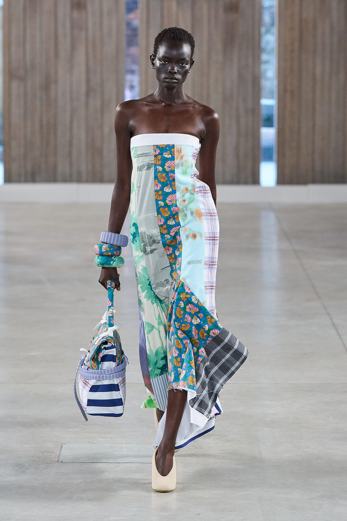 Model walking in a strapless patchwork dress with floral and plaid panels, carrying a striped bag and wearing chunky bracelets.