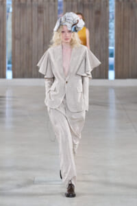 Model walks the runway in a light beige oversized suit with dramatic cape-like shoulders and a striped head wrap.