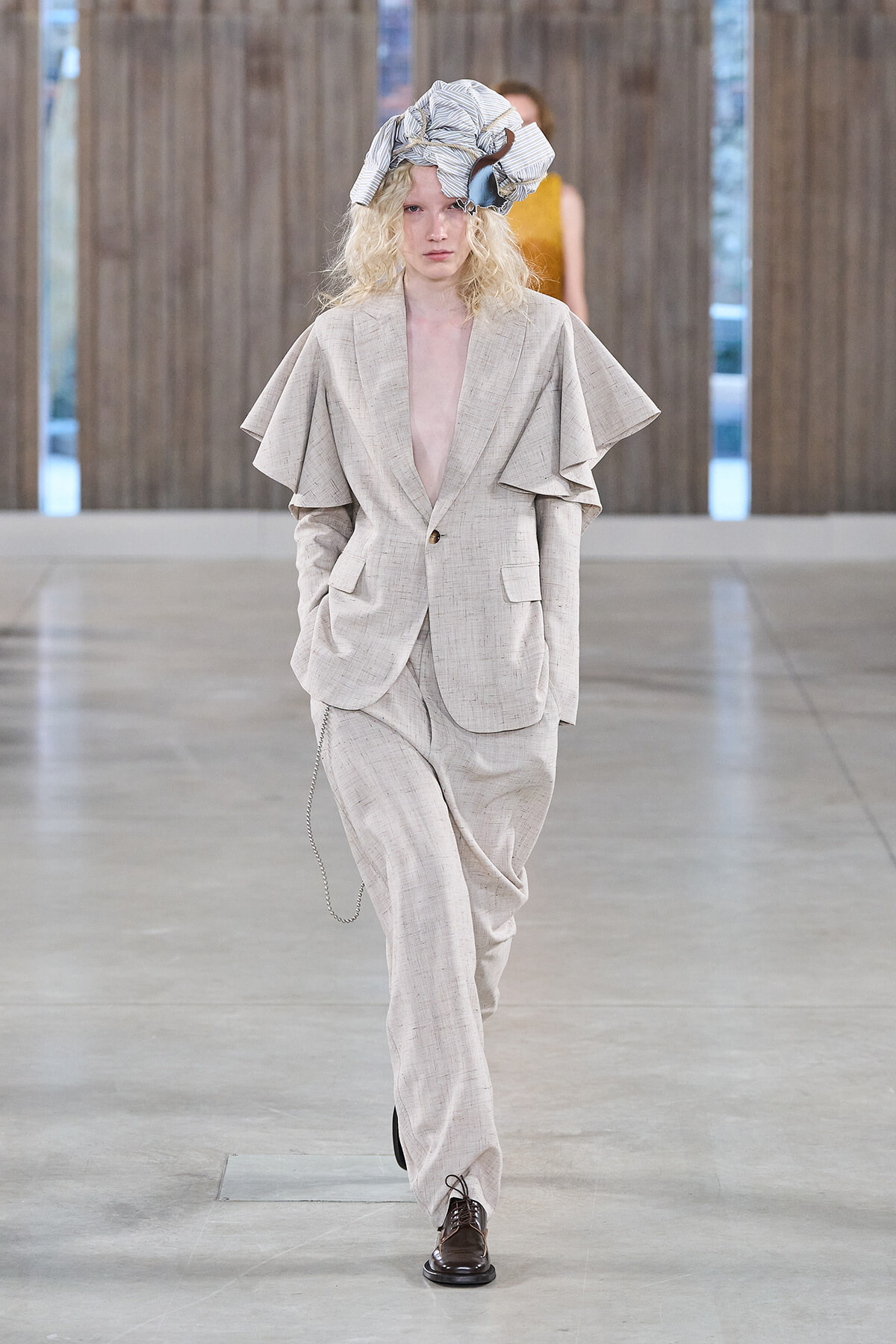 Model walks the runway in a light beige oversized suit with dramatic cape-like shoulders and a striped head wrap.