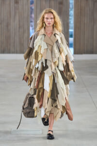Model walking in a beige-toned patchwork dress made of fabric scraps, carrying a brown backpack.