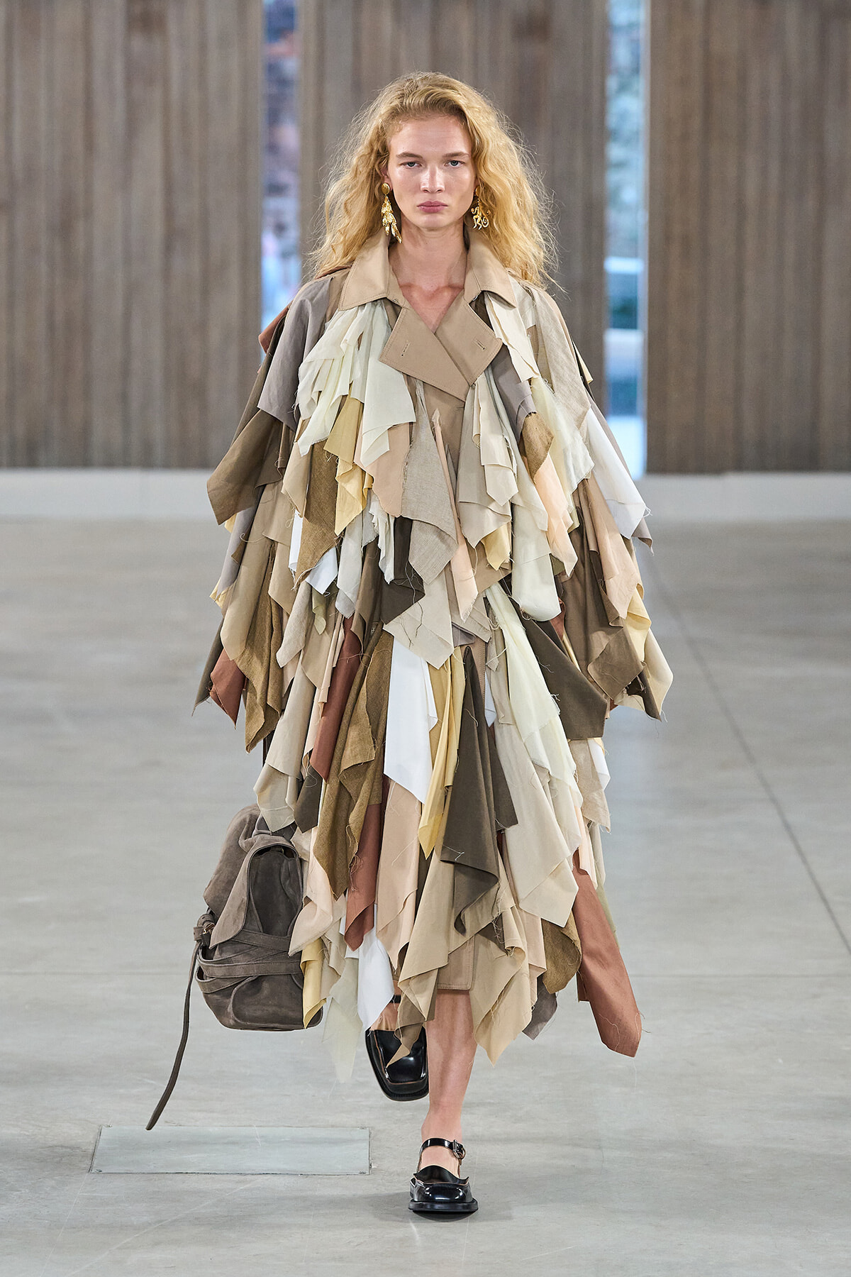 Model walking in a beige-toned patchwork dress made of fabric scraps, carrying a brown backpack.
