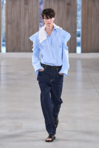 Male model walking a runway in a light blue button-down shirt with a white ruffled accessory and dark jeans, hands in pockets.
