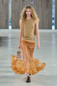 Model walking runway in tan sleeveless top and layered sheer orange skirt, carrying a Moschino-brand bag and wearing brown shoes.