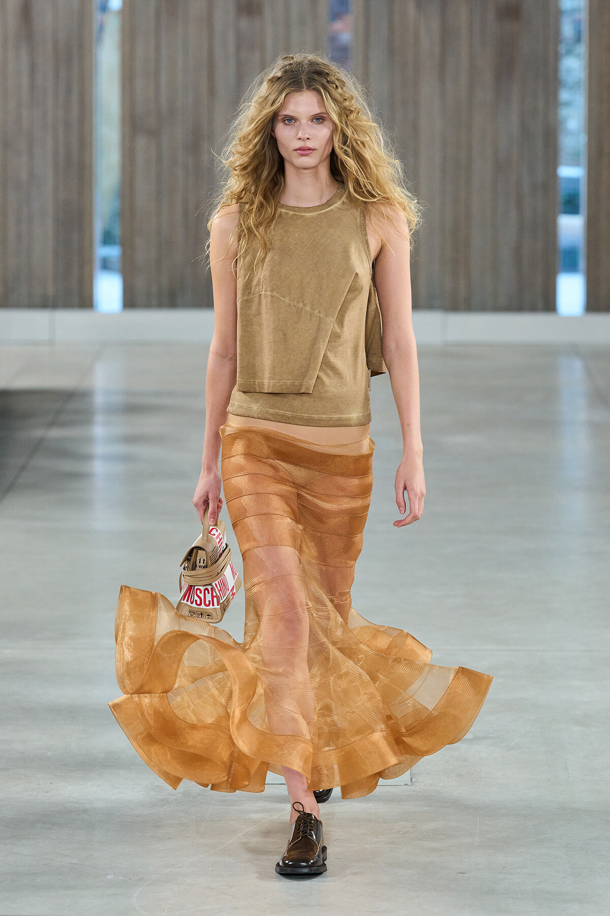 Model walking runway in tan sleeveless top and layered sheer orange skirt, carrying a Moschino-brand bag and wearing brown shoes.