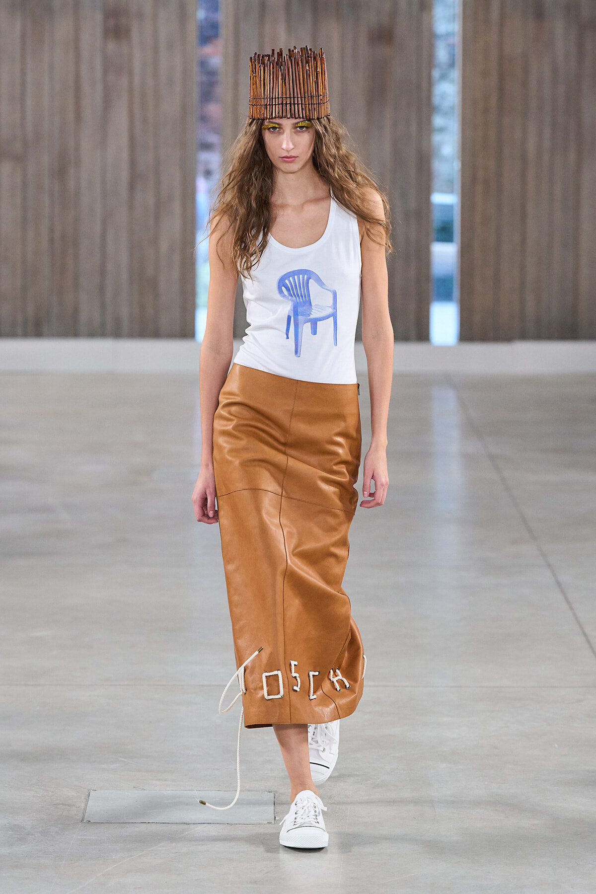 Model on a runway wearing a white tank with a blue chair graphic, tan leather midi skirt with rope details, white sneakers, and a tall wooden stick headdress.