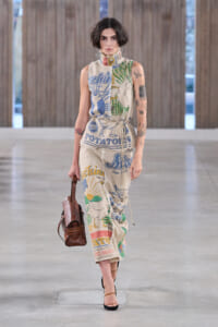 Model walking a runway in a sleeveless, oversized jumpsuit with bold, colorful graphic prints and text (potato and sun motifs); brown handbag in left hand; brown heeled sandals; short dark hair.