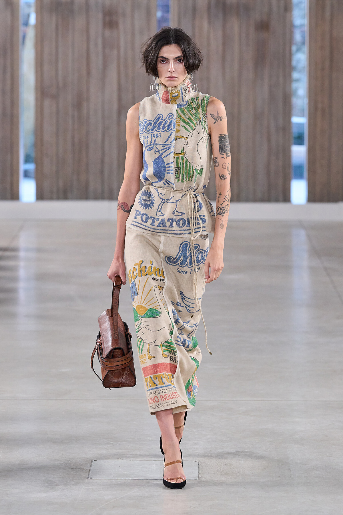 Model walking a runway in a sleeveless, oversized jumpsuit with bold, colorful graphic prints and text (potato and sun motifs); brown handbag in left hand; brown heeled sandals; short dark hair.