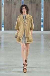 Model on a runway wearing a tan oversized tunic with lace-up front, gray inner dress, and large cargo pockets, brown platform sandals.