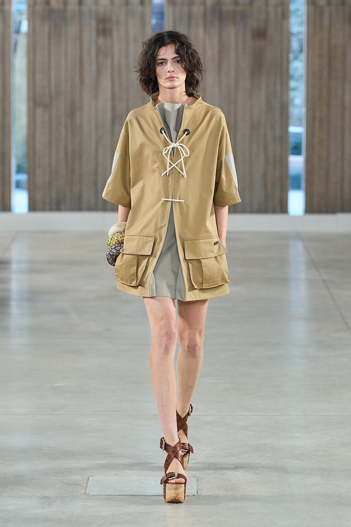 Model on a runway wearing a tan oversized tunic with lace-up front, gray inner dress, and large cargo pockets, brown platform sandals.