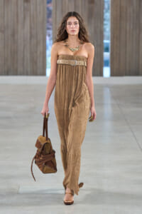 Model walks a runway in a strapless tan pleated jumpsuit with a wide beige belt and layered necklace, carrying a brown handbag.