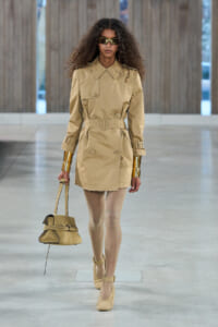 Female model walking a runway in a tan trench-style dress with belt, gold cuffs, a matching handbag, and beige heels with sunglasses nearby her face.
