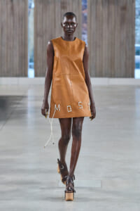 Model walks the runway wearing a sleeveless tan leather mini dress with white stitched letters along the hem and white cords hanging from the side.