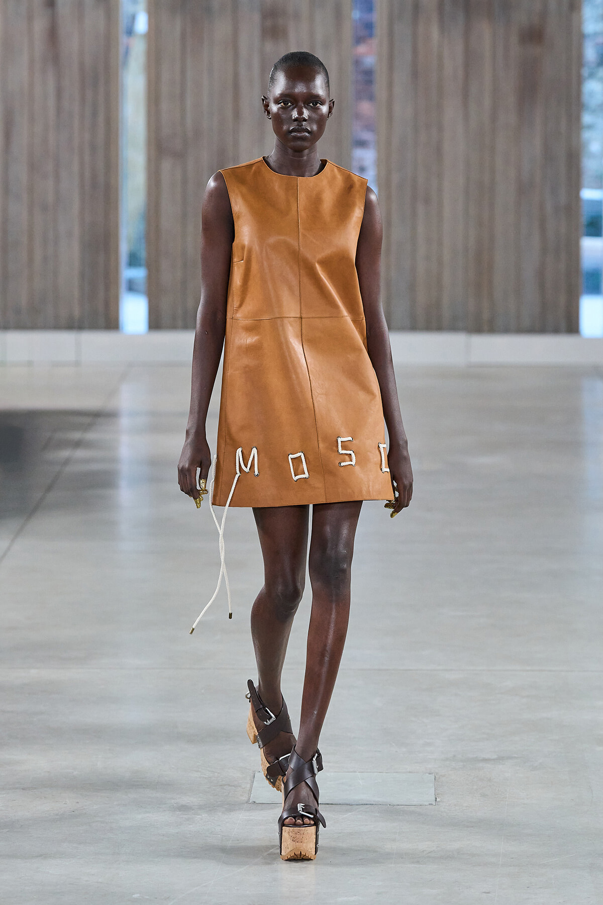 Model walks the runway wearing a sleeveless tan leather mini dress with white stitched letters along the hem and white cords hanging from the side.