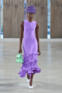 Model on a runway wearing a lavender sleeveless dress with a large ruffled hem, purple head wrap, white heels, and a mint-green handbag.