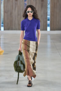 Runway model wearing a cobalt purple polo and color-block striped wide-leg pants, with white sunglasses and a green studded bag.