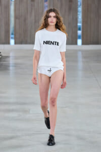 Model walking a fashion runway in an all-white outfit: oversized T-shirt with 'NIENTE' and white briefs with a small black graphic, black shoes.