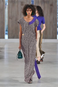 Model walking a runway in a long black-and-white floral maxi dress with a purple neckline panel, green earrings, and a small green handbag edge.