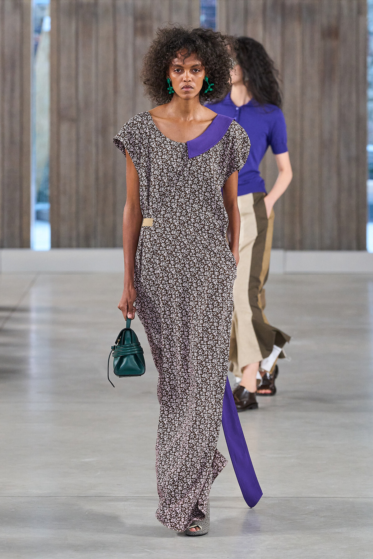 Model walking a runway in a long black-and-white floral maxi dress with a purple neckline panel, green earrings, and a small green handbag edge.