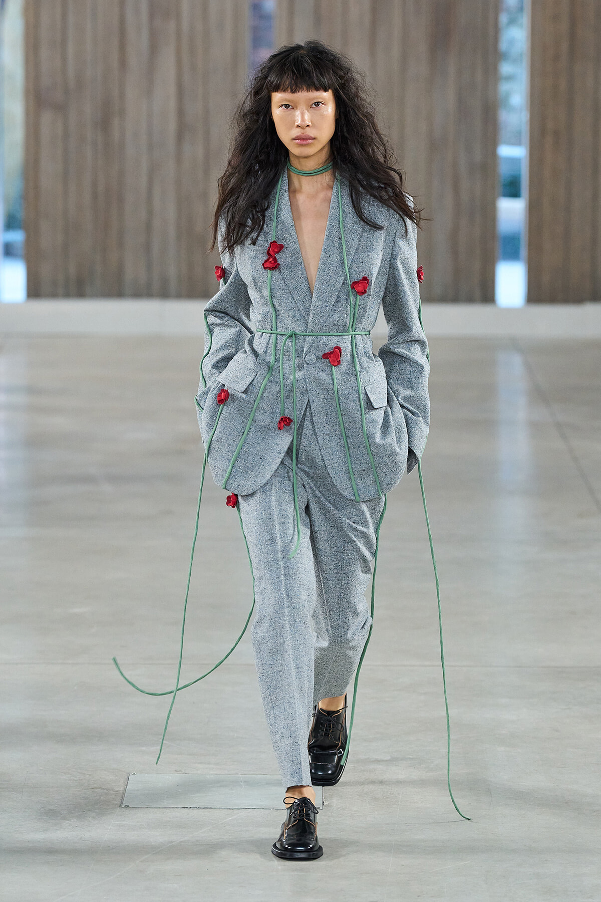 Model walking a runway in a light gray suit with green cords and red floral appliqués, indoors at a fashion show.