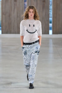 Female fashion model walking a runway in a white knit sweater featuring a black smiley face and newspaper-print pants with a black belt.